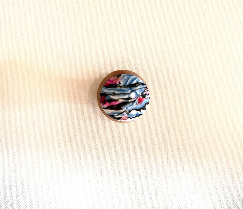 K&ouml;nnte beinhalten: Eine kleine, runde Holzkiste mit einem farbenfrohen, abstrakten Design auf dem Deckel. Das Design zeigt Wirbel in Blau, Rosa und Schwarz.