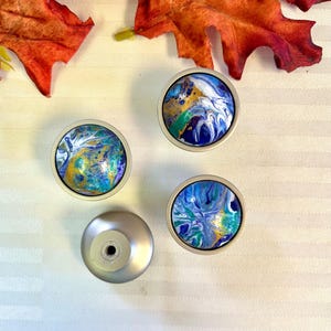 May include: Four decorative cabinet knobs with abstract art designs. The knobs feature a silver base and colorful, swirling patterns in shades of blue, gold, and green. The knobs are arranged on a white striped surface.