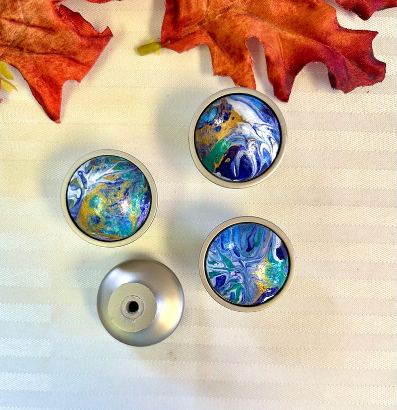 May include: Four decorative cabinet knobs with abstract art designs. The knobs feature a silver base and colorful, swirling patterns in shades of blue, gold, and green. The knobs are arranged on a white striped surface.