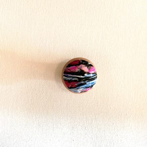 K&ouml;nnte beinhalten: Ein runder Holz-Knopf mit einem farbenfrohen abstrakten Design. Das Design zeigt Wirbel in Pink, Schwarz, Wei&szlig; und Blau.