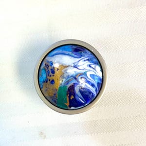 May include: A round cabinet knob with a silver metal base. The knob's face features a swirling design in shades of blue, white, gold, and green, resembling an abstract painting. The knob is designed for furniture and home decor.