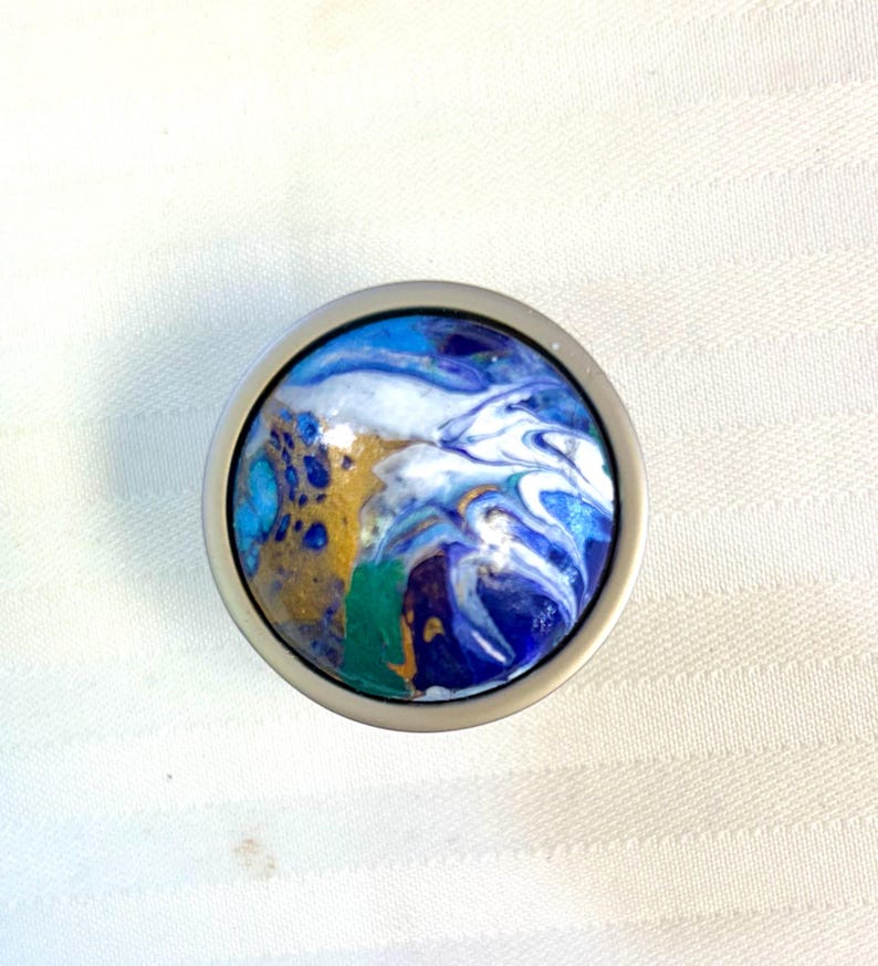 May include: A round cabinet knob with a silver metal base. The knob's face features a swirling design in shades of blue, white, gold, and green, resembling an abstract painting. The knob is designed for furniture and home decor.