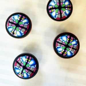 May include: Four decorative cabinet knobs with a colorful, mosaic-like design. Each knob features a black background with blue, pink, green, and white patterns, encased in a dark bronze-colored metal frame. The knobs are arranged on a white, textured surface.
