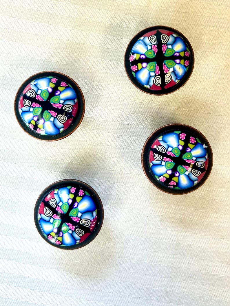 May include: Four decorative cabinet knobs with a mosaic design. Each knob has a black background with blue, pink, green, and white patterns, set in a dark bronze-coloured metal frame. The knobs are arranged on a white, textured surface.
