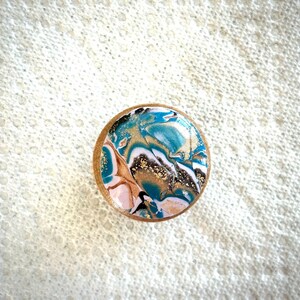 May include: A round, decorative wooden knob with a marbled design. The design features swirls of teal, gold, white, black, and pink. The knob has a light brown wooden edge and is set against a textured white background.
