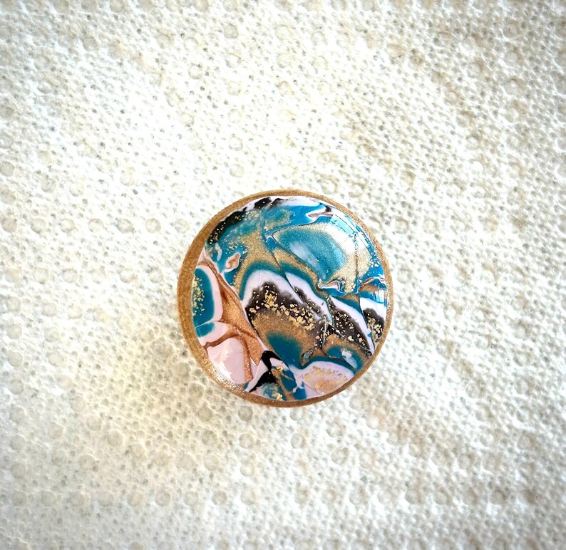 May include: A round, decorative wooden knob with a marbled design. The design features swirls of teal, gold, white, black, and pink. The knob has a light brown wooden edge and is set against a textured white background.