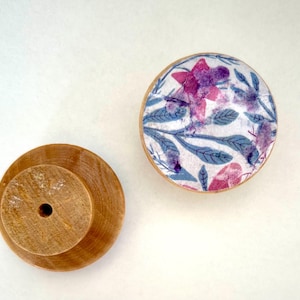 K&ouml;nnte beinhalten: Zwei Holzschrankkn&ouml;pfe. Einer ist unbehandelt, mit zylindrischem Sockel und zentralem Loch. Der andere ist rund, mit einem floralen Design in Rosa und Lila auf wei&szlig;em Hintergrund, umrahmt von einem hellen Holzrand. Das florale Design umfasst blaue Bl&auml;tter.