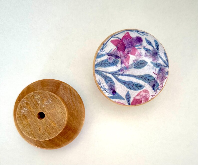 K&ouml;nnte beinhalten: Zwei Holzschrankkn&ouml;pfe. Einer ist unbehandelt, mit zylindrischem Sockel und zentralem Loch. Der andere ist rund, mit einem floralen Design in Rosa und Lila auf wei&szlig;em Hintergrund, umrahmt von einem hellen Holzrand. Das florale Design umfasst blaue Bl&auml;tter.