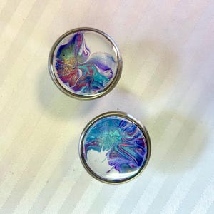 May include: Two round, silver-coloured knobs with a white background and abstract swirls of purple, blue, and pink paint.