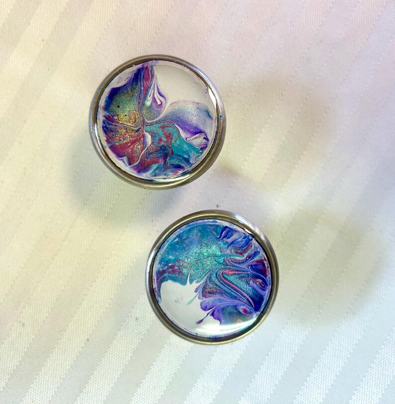 May include: Two round, silver-coloured knobs with a white background and abstract swirls of purple, blue, and pink paint.