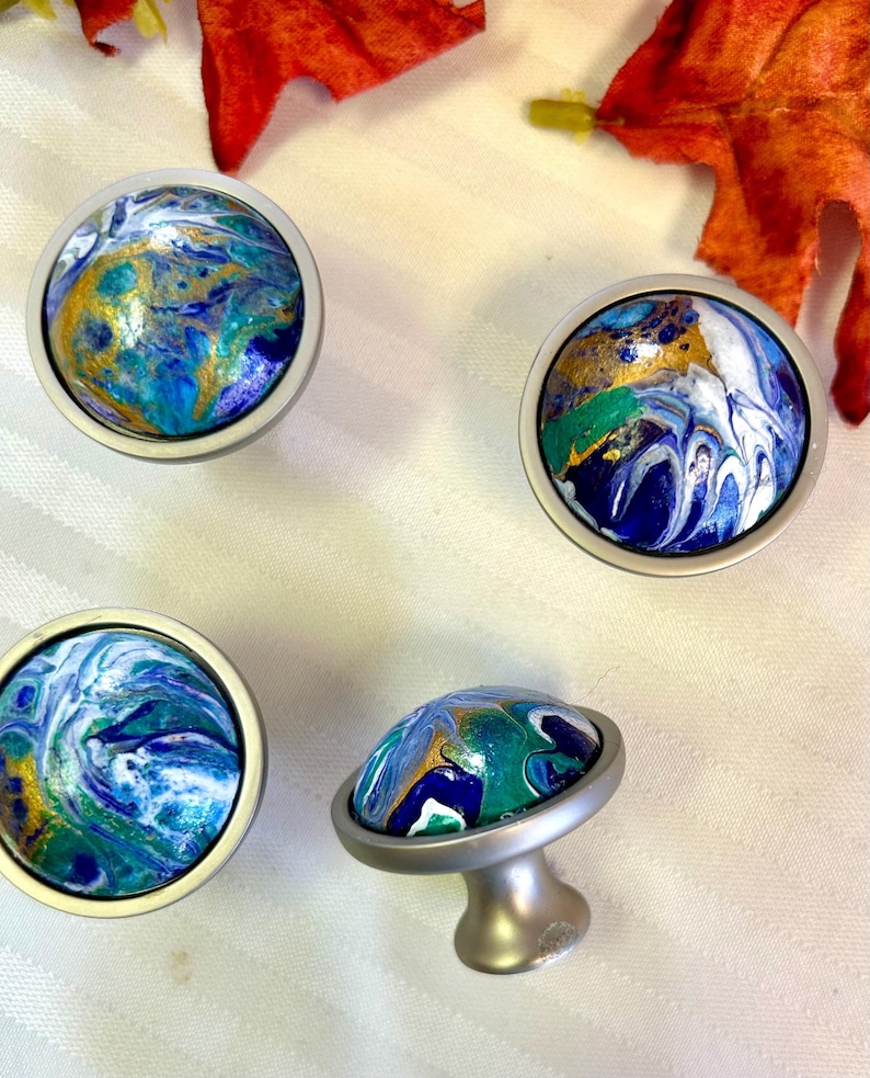May include: Four cabinet knobs with a unique, abstract design. Each knob features a round, silver-toned base and a colorful, swirled pattern in shades of blue, green, gold, and white. One knob is positioned upright, showcasing its mushroom-shaped stem.