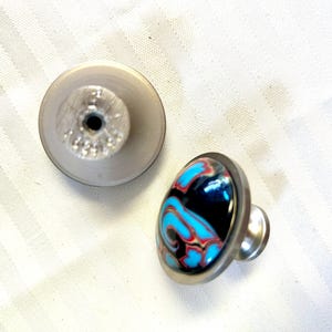 May include: Two cabinet knobs, one with a silver base and a blue, black, and red marbled top, and the other with a silver base and a plain silver top.