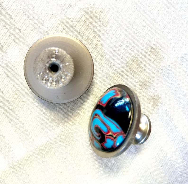 May include: Two cabinet knobs, one with a silver base and a blue, black, and red marbled top, and the other with a silver base and a plain silver top.