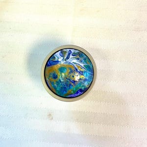 May include: A round, silver-toned knob with a colorful, abstract design. The artwork features swirls of blue, teal, gold, and white, creating a marbled effect. The knob is likely for a cabinet or drawer.