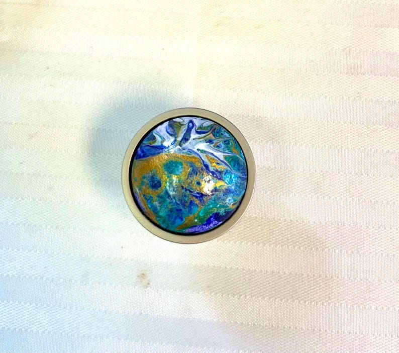 May include: A round, silver-toned knob with a colorful, abstract design. The artwork features swirls of blue, teal, gold, and white, creating a marbled effect. The knob is likely for a cabinet or drawer.