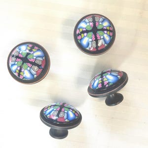 May include: Four decorative cabinet knobs with a colourful, mosaic-like design. The knobs feature a black base with a bronze-coloured rim and a patterned top with blue, pink, green, and white elements. The knobs are round and have a small, dark stem.