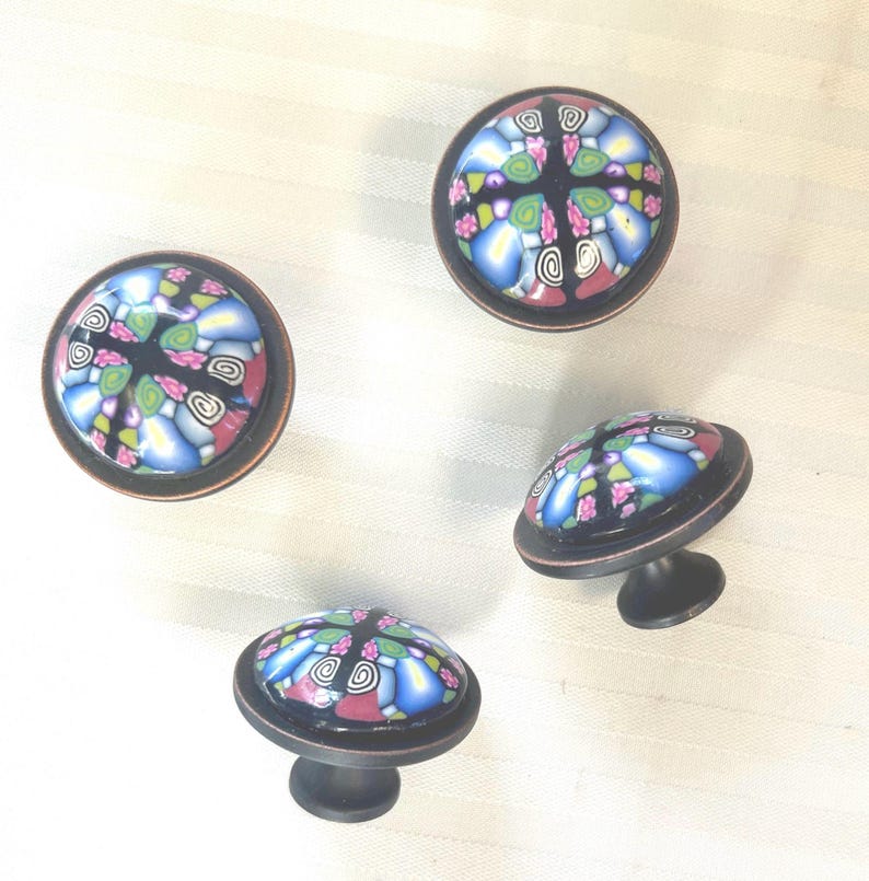 May include: Four decorative cabinet knobs with a colourful, mosaic-like design. The knobs feature a black base with a bronze-coloured rim and a patterned top with blue, pink, green, and white elements. The knobs are round and have a small, dark stem.