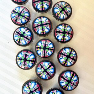 May include: A set of decorative cabinet knobs. Each knob has a colourful, mosaic-style design with blue, pink, green, and yellow patterns on a black background. The knobs have a copper-coloured metal base.