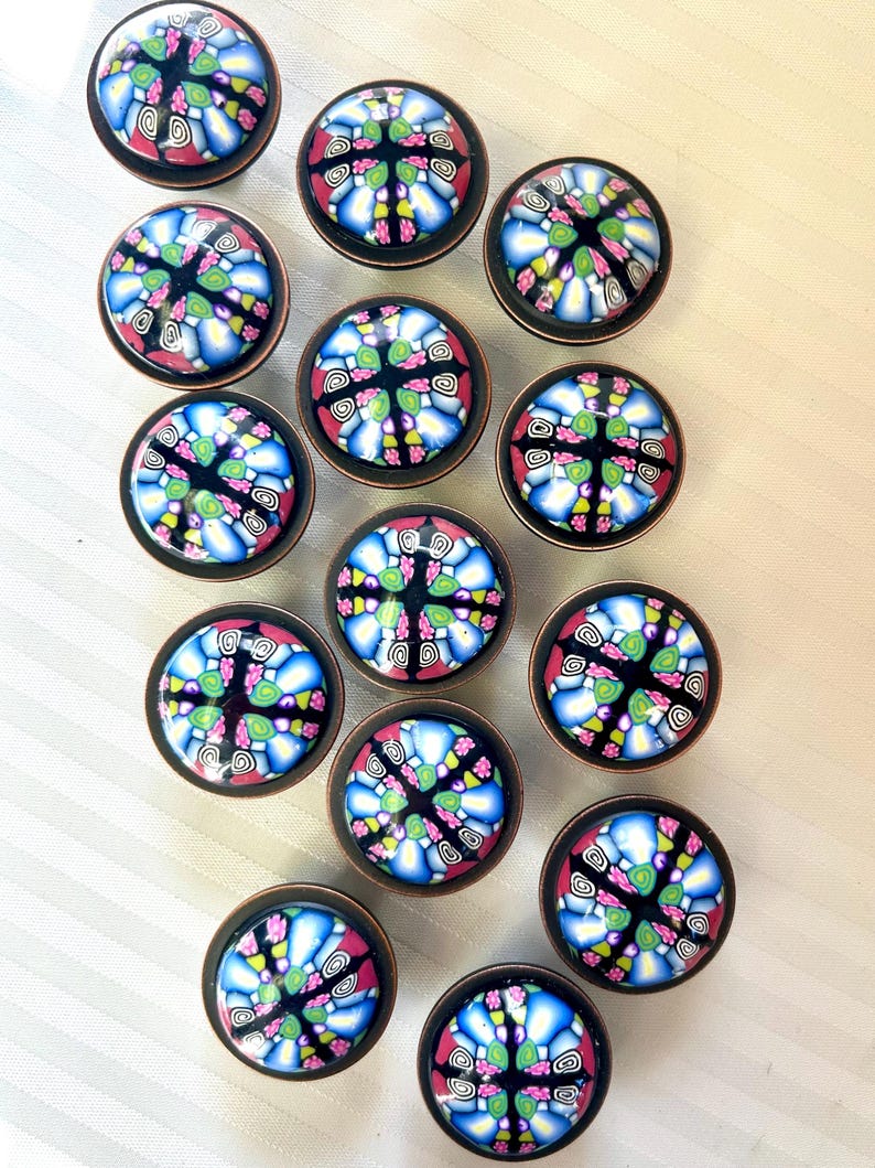 May include: A set of decorative cabinet knobs. Each knob has a colourful, mosaic-style design with blue, pink, green, and yellow patterns on a black background. The knobs have a copper-coloured metal base.