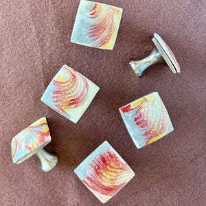 May include: Five square cabinet knobs with a marbled design in shades of red, yellow, and white. Two of the knobs are attached to silver bases.