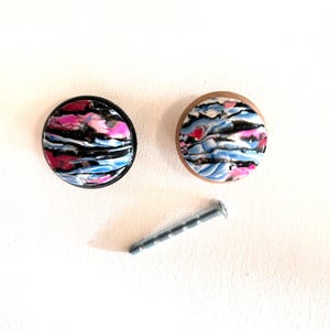 K&ouml;nnte beinhalten: Zwei runde Schrankkn&ouml;pfe mit schwarzem und h&ouml;lzernem Sockel. Die Kn&ouml;pfe haben ein farbenfrohes abstraktes Design mit rosa, blauen und schwarzen Wirbeln.