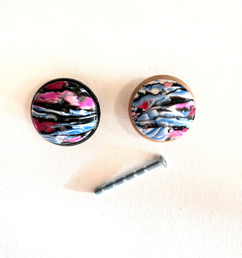 K&ouml;nnte beinhalten: Zwei runde Schrankkn&ouml;pfe mit schwarzem und h&ouml;lzernem Sockel. Die Kn&ouml;pfe haben ein farbenfrohes abstraktes Design mit rosa, blauen und schwarzen Wirbeln.