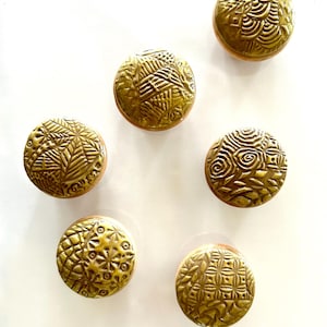 May include: Six round, gold-colored ceramic knobs with different embossed patterns. The knobs have a slightly raised edge around the perimeter.