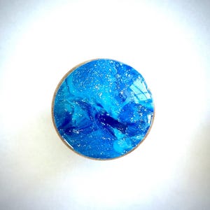 K&ouml;nnte beinhalten: Ein runder Flaschenverschluss aus Holz mit einem abstrakten blau-wei&szlig;en Design. Die Oberseite hat eine gl&auml;nzende Oberfl&auml;che mit wirbelnden Mustern und gesprenkelten Akzenten, die einem Ozean &auml;hneln. Durchmesser ca. 3 cm.