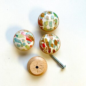 May include: Four round wooden knobs with a colourful abstract design. The knobs have a white background with splashes of red, blue, green, and gold paint. One knob is shown from the bottom, revealing a screw hole.