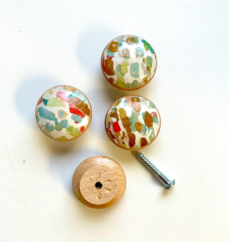 May include: Four round wooden knobs with a colourful abstract design. The knobs have a white background with splashes of red, blue, green, and gold paint. One knob is shown from the bottom, revealing a screw hole.