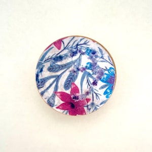 K&ouml;nnte beinhalten: Ein rundes Objekt mit floralem Muster und Holzrand. Das Design zeigt einen wei&szlig;en Hintergrund mit blauen, lila und pinkfarbenen Blumen und Bl&auml;ttern. Das Objekt ist vor einem wei&szlig;en Hintergrund zentriert.