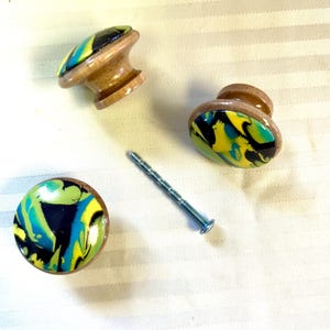 May include: Three wooden cabinet knobs with colorful, swirled epoxy resin tops in shades of yellow, blue, green, and black. A silver screw is also present. The knobs have a natural wood base.