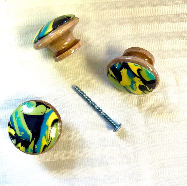 May include: Three wooden cabinet knobs with colorful, swirled epoxy resin tops in shades of yellow, blue, green, and black. A silver screw is also present. The knobs have a natural wood base.