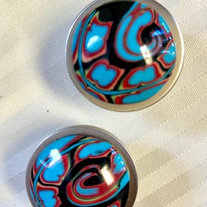 May include: A pair of round, silver-toned earrings with a black, blue, red, and green swirl pattern.