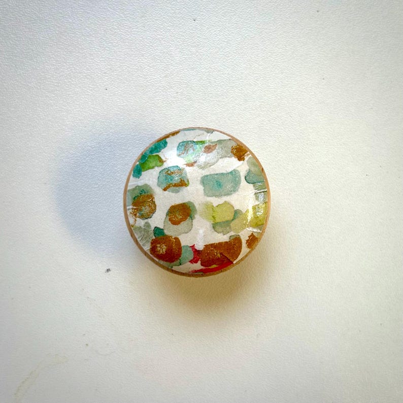 May include: A round, wooden knob with a colourful abstract design. The design features shades of green, blue, yellow, orange, and red.