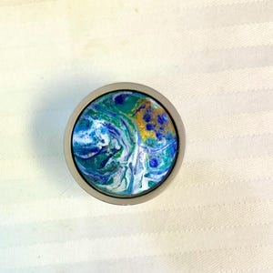 May include: A round cabinet knob with a swirling design in shades of blue, green, white, and gold. The knob is set in a silver-colored metal frame. The design resembles a miniature abstract painting.