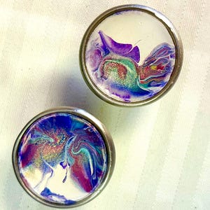 May include: Two round, silver-coloured drawer knobs with a white background and abstract purple, blue, and green paint swirls. The paint has a glittery finish.