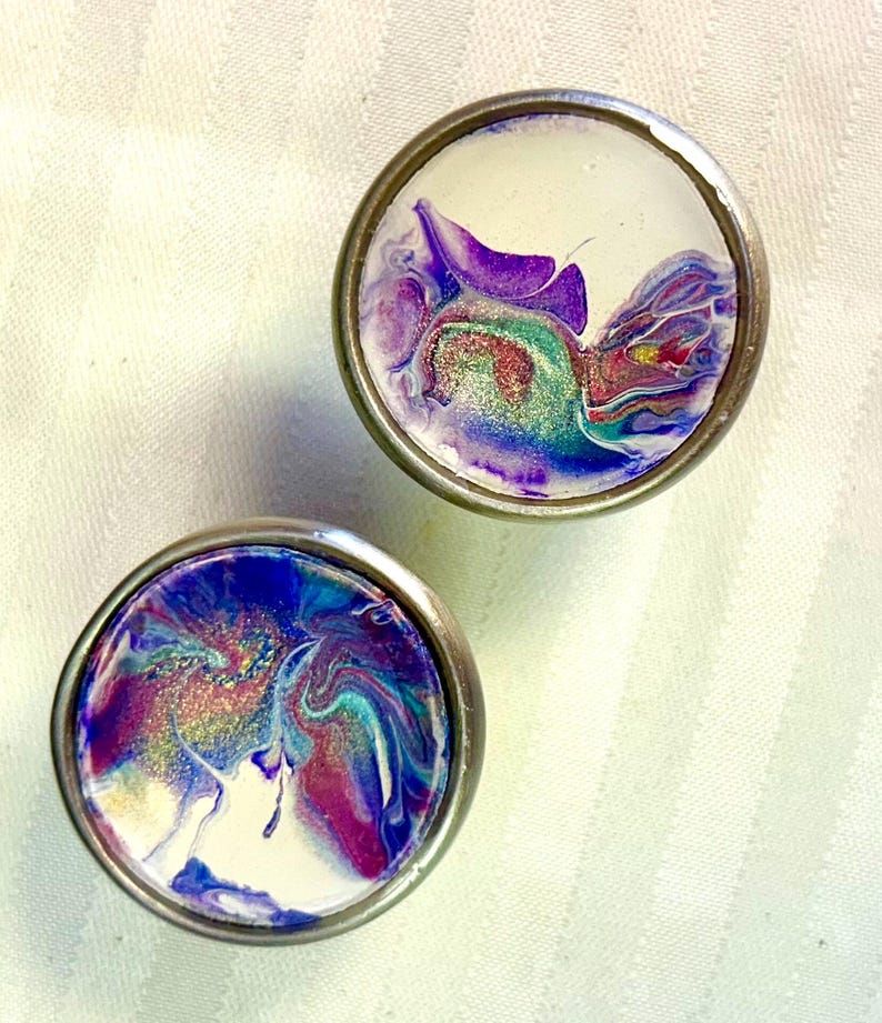 May include: Two round, silver-coloured drawer knobs with a white background and abstract purple, blue, and green paint swirls. The paint has a glittery finish.
