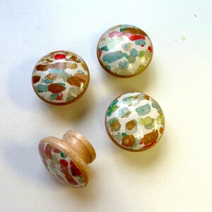 May include: Four wooden knobs with a white background and colourful abstract patterns. The knobs are round and have a raised edge. The patterns are made up of red, orange, yellow, green, blue, and purple.