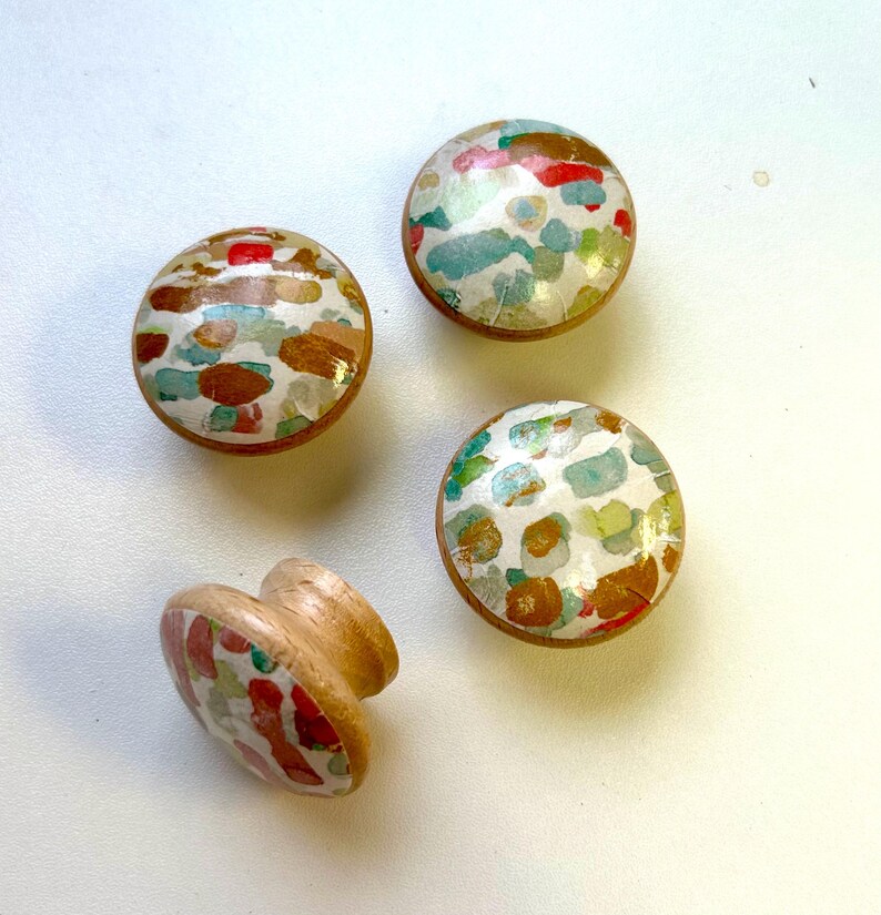 May include: Four wooden knobs with a white background and colourful abstract patterns. The knobs are round and have a raised edge. The patterns are made up of red, orange, yellow, green, blue, and purple.
