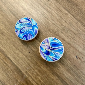 May include: Two round, blue and white decorative objects with a floral design. The design features shades of blue and purple with gold accents. The objects are set against a wooden background.