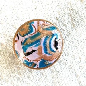 May include: A round, decorative object with a light pink, teal, gold, and black abstract design. The edges are a light brown color. The object has a glossy finish and appears to be made of resin or a similar material.