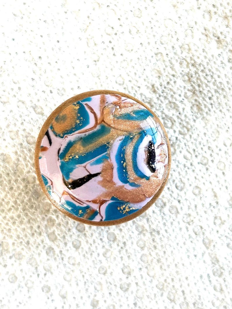 May include: A round, decorative object with a light pink, teal, gold, and black abstract design. The edges are a light brown color. The object has a glossy finish and appears to be made of resin or a similar material.