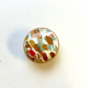 May include: A round wooden knob with a white background and abstract pattern in shades of brown, gold, green, and red.