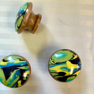 May include: Three wooden cabinet knobs with a colorful, swirled design in shades of yellow, blue, green, and black. The knobs have a round, flat face and a wooden base.