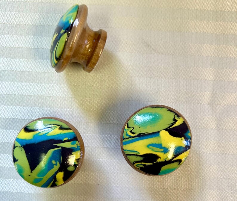 May include: Three wooden cabinet knobs with a colorful, swirled design in shades of yellow, blue, green, and black. The knobs have a round, flat face and a wooden base.