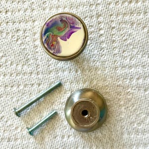 May include: A set of two silver cabinet knobs with screws. One knob features a white background with a colourful abstract design in shades of purple, blue, green, and red.