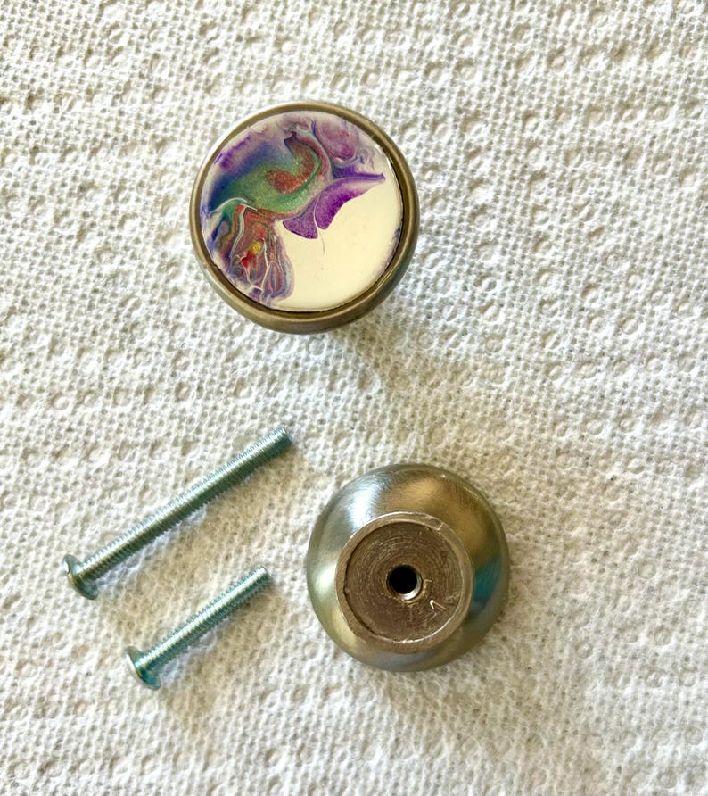 May include: A set of two silver cabinet knobs with screws. One knob features a white background with a colourful abstract design in shades of purple, blue, green, and red.