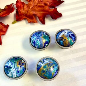 May include: Four round cabinet knobs with a silver finish. The knobs feature a colorful, abstract design in shades of blue, green, and gold. The knobs are arranged on a white surface with red and orange fall leaves.
