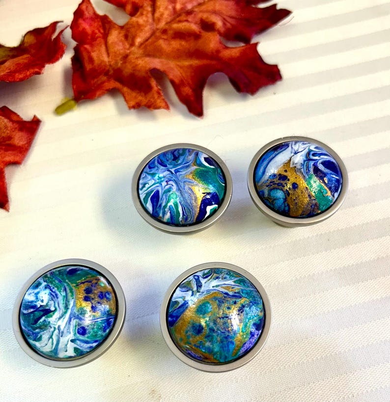 May include: Four round cabinet knobs with a silver finish. The knobs feature a colorful, abstract design in shades of blue, green, and gold. The knobs are arranged on a white surface with red and orange fall leaves.